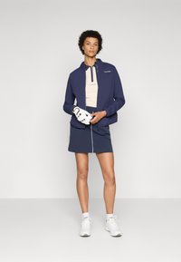 Navy windbreaker jacket, beige polo shirt, navy skirt with white zipper detail. White athletic shoes and gloves complete the outfit.