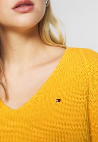 Żółty sweter dziany z dekoltem w kształcie litery V. Ma małe logo w kolorach czerwonym, białym i niebieskim po lewej stronie, blisko ramienia.