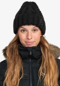 Black knitted beanie with textured cable design. Worn with a black jacket featuring a faux fur collar. Soft, warm material.