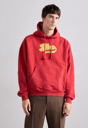 Hoodie en coton rouge avec cordons de serrage, arborant un logo "Blue" jaune et une poche avant. Porté avec un pantalon marron.