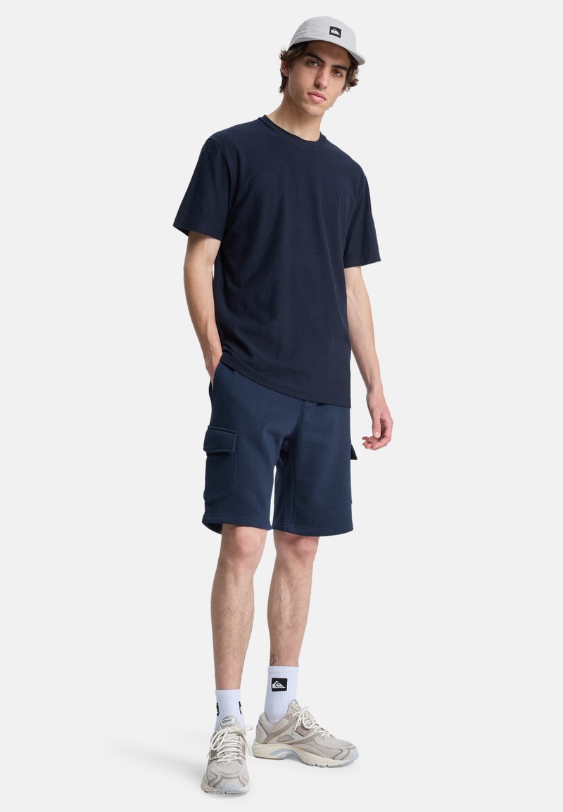Navy blue cotton t-shirt with short sleeves, paired with navy cargo shorts featuring side pockets. Light gray cap and cream sneakers.