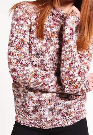 Femme portant un pull en grosse maille multicolore avec des nuances de blanc, rose, orange et marron, bras croisés, avec de longs cheveux auburn.