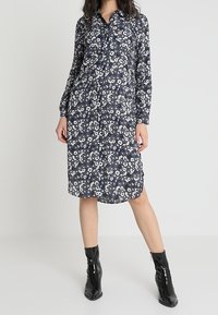 Blommig skjortklänning i marinblått med vita blommor; långärmad, med krage, avslappnad passform, medellång, tillsammans med svarta ankelstövlar.