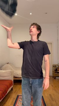 Νέος άνδρας με μαύρη μπλούζα και τζιν πετάει ένα μαύρο σακάκι σε ένα απλό δωμάτιο με ξύλινο πάτωμα και έπιπλα.