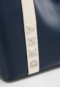 Marinblå läderhandväska med en krämfärgad rem med metallisk DKNY-tryck. Slät textur och strukturerad form med rena linjer.