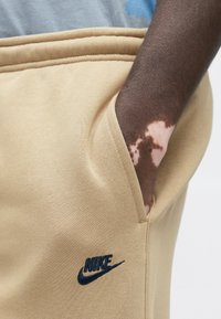 Bézs színű melegítőnadrág puha anyaggal, oldalzsebbel és tengerészkék Nike logóval. Az anyag sima és enyhén rugalmas.