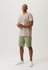 Beżowy t-shirt z grafikiem i napisem "Diverse", w połączeniu z zielonymi szortami cargo i białymi sneakersami. Prosty, casualowy wygląd.