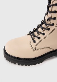 Bottines de combat en cuir beige avec des coutures noires, une semelle épaisse noire et des lacets noirs passés à travers des œillets en métal.
