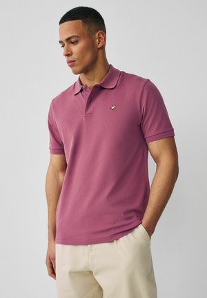 Man draagt een mauve polo met een klein vogel-logo op de borst, beige broek, staat met één hand in de zak, kijkt naar beneden links.