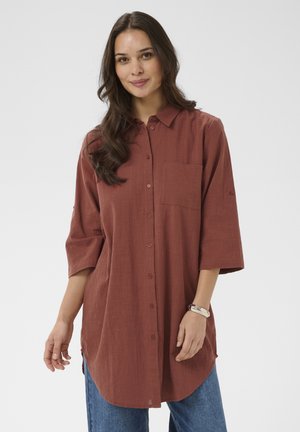 Žena s dlhými tmavými vlasmi, oblečená v voľnej hrdzavej košeli na gombíky a modrých džínsoch, stojaca pred jednofarebným svetlým pozadím.