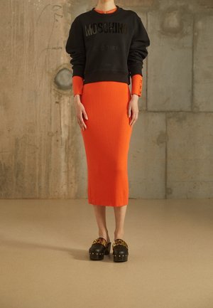 Personne portant un sweat-shirt noir Moschino sur une longue robe orange côtelée et des chaussures compensées noires avec logo Moschino doré.