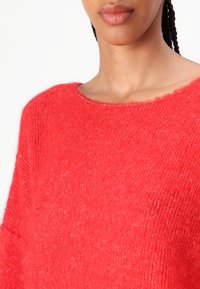 Pull rouge léger avec une coupe ample, un col large et une texture douce. Présente un ourlet légèrement effiloché au niveau du col.