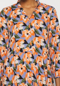 Robe florale à manches trois-quarts, ornée de fleurs orange et blanches sur un fond bleu clair. Col froncé et détails de boutons.