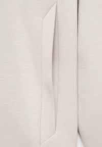 Un țesut textil de culoare bej deschis, cu textură, care prezintă o buzunare lateral cu o cusătură curată. Materialul are un finisaj moale, ușor melanjat.