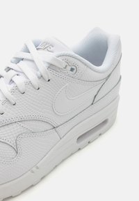 Biele tenisky Nike s textúrovaným koženým zvrškom, vypolstrovaným golierom a viditeľnou vzduchovou jednotkou; majú biele šnúrky a branding na jazyku.