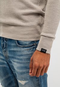 Helles graues, strukturiertes Strickoberteil mit gerippten Bündchen, kombiniert mit ausgewaschenen blauen Jeans. Das Etikett auf dem Bündchen zeigt "J BUTCHER OF BLUE".