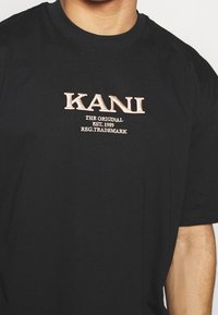 Černé bavlněné tričko se vyšívaným textem "KANI" v světle béžové barvě, pod nímž je v menším písmu "THE ORIGINAL EST. 1989 REG. TRADEMARK".