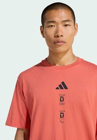 Camiseta de algodón color naranja coral con cuello redondo, mangas cortas y logo impreso en negro de Adidas con gráficos del equipo olímpico y paralímpico.