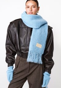 Šviesiai mėlyna banguota šalikėlis, pagamintas iš minkštos vilnos, su kutais gale, derinamas su tamsiai ruda odine striuke ir atitinkančiais šviesiai mėlynais pirštinėmis.