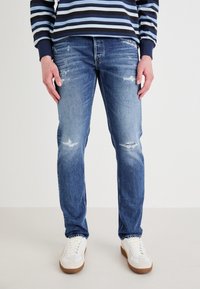 Jeans de mezclilla azul claro con un corte ajustado, que presentan desgastes y desgarros. Combinados con zapatillas blancas y una camisa de manga larga a rayas azul marino.
