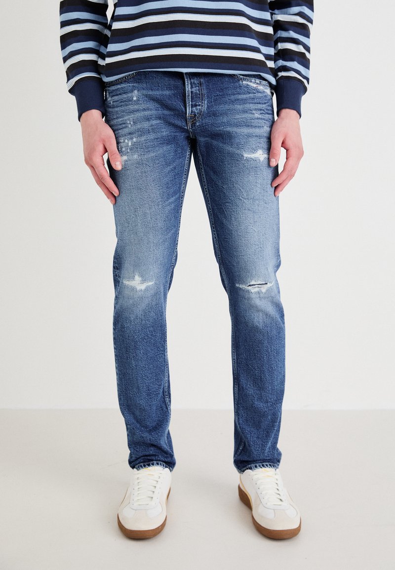 Jeans de mezclilla azul claro con un corte ajustado, que presentan desgastes y desgarros. Combinados con zapatillas blancas y una camisa de manga larga a rayas azul marino.