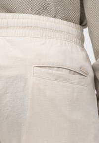 Svijetlosmeđi hlače s teksturiranom tkaninom, elastičnim pojasom i malim detaljem stražnjeg džepa. Jednostavan, čist dizajn.