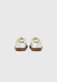 Ζευγάρι λευκά αθλητικά παπούτσια με καφέ σόλες, εμφανισμένα από πίσω, με την εμπορική επωνυμία «VEJA» σε μαύρο χρώμα σε κάθε ταμπέλα της φτέρνας.