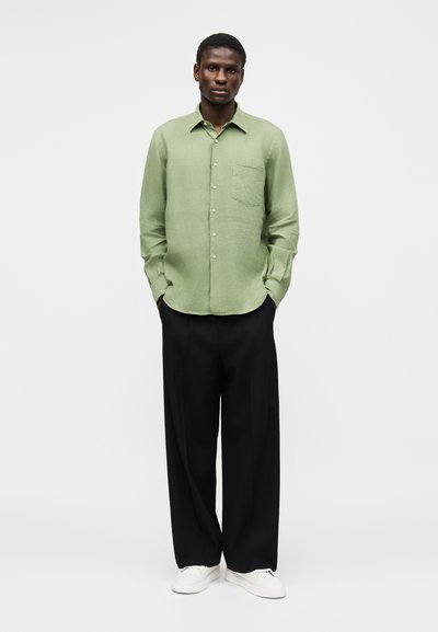 Άνδρας στέκεται με τα χέρια στις τσέπες, φοράει ένα ανοιχτοπράσινο πουκάμισο με κουμπιά, μαύρο παντελόνι με φαρδιά μπατζάκια και λευκά αθλητικά παπούτσια σε απλό φόντο.