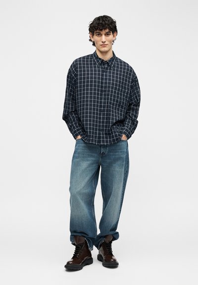 Νέος άνδρας που στέκεται με τα χέρια στις τσέπες, φοράει πουκάμισο με ασπρόμαυρο καρό, φαρδιά μπλε τζιν και σκούρα καφέ μπότες με κορδόνια.