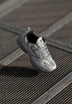 Gray and white FILA sneaker with black accents, placed on an industrial metal grate floor in soft lighting.