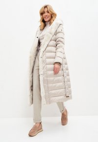 Abrigo puff largo beige con cuello de piel sintética, que presenta un acolchado horizontal, bolsillos laterales y cierre de botones, combinado con pantalones de color claro.