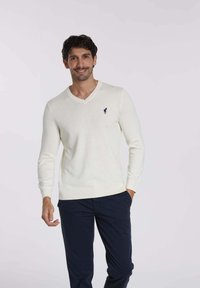 Pull crème en V fabriqué dans un tissu doux, avec des manches longues et un logo marine sur la poitrine. Associé à un pantalon marine.