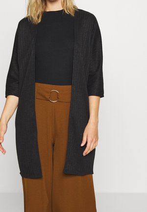 Kobieta ubrana w czarny prążkowany otwarty kardigan, czarny top oraz brązowe spodnie o szerokich nogawkach i wysokim stanem, z okrągłą klamrą paska.