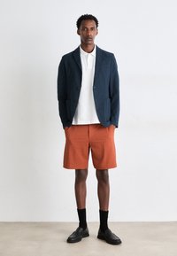 Sacou texturat de culoare bleumarin, tricou polo alb, pantaloni scurți de culoare roșie cu un model subtil, șosete negre și pantofi slip-on negri.