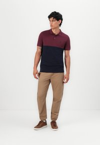 Miestenska polo košeľa so vínovým horným dielom a námornícky modrým spodným dielom, vyrobená zo 100% bavlny, s logom na hrudi. Skombinovaná s khaki nohavicami a hnedými topánkami.