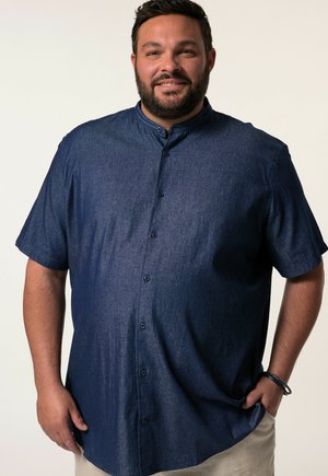 Homme barbu portant une chemise bleu foncé à manches courtes boutonnée, debout avec une main dans la poche sur un fond uni.