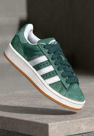 Grüner Wildledersneaker mit weißen Streifen, dunkelgrünen Schnürsenkeln und weißer Sohle mit Gummiboden, auf einer Betonfläche positioniert.