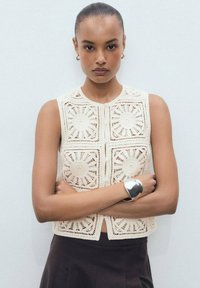 Beige crochet crop top featuring circular pattern detailing, square design, round neckline, and front button closure, paired with a dark skirt.