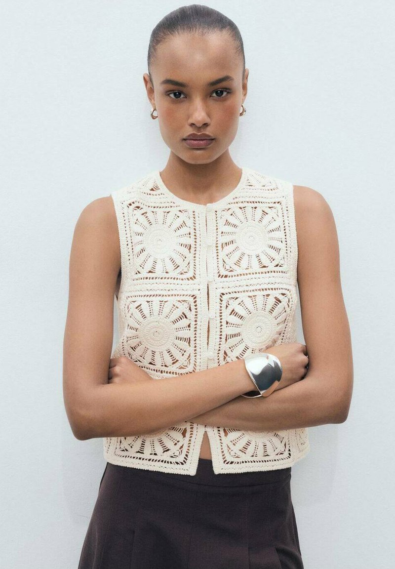 Beige crochet crop top featuring circular pattern detailing, square design, round neckline, and front button closure, paired with a dark skirt.