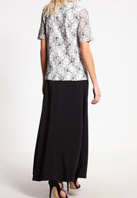 Top de encaje con mangas cortas, patrón blanco y negro, combinado con una falda maxi negra. El conjunto presenta una silueta fluida y sandalias planas.