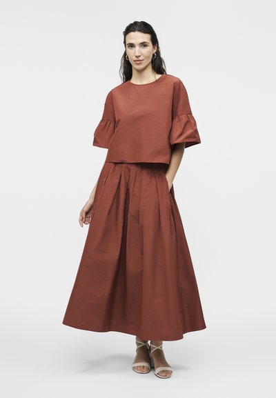 Γυναίκα που φοράει καφέ-σκουρόχρωμο φαρδύ τοπ με φαρδιά μανίκια και μακριά πλισέ φούστα στον ίδιο τόνο, στέκεται με το ένα χέρι στην τσέπη, λευκά σανδάλια.