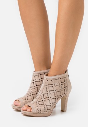Bottines en suède beige à talons hauts, avec des motifs géométriques découpés, des orteils ouverts et une fermeture éclair à l'arrière, sur les jambes d'une personne.