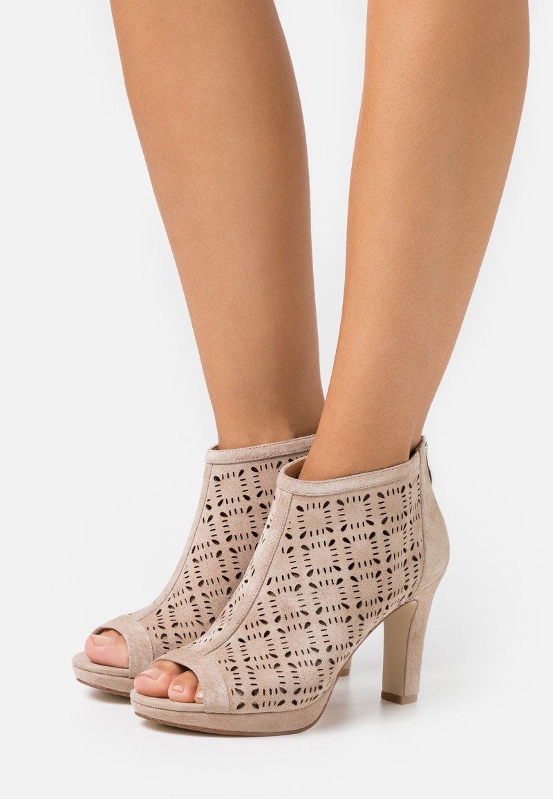 Botines de tacón alto de ante beige con patrones geométricos recortados, punta abierta y una cremallera en la parte trasera, sobre las piernas de una persona.