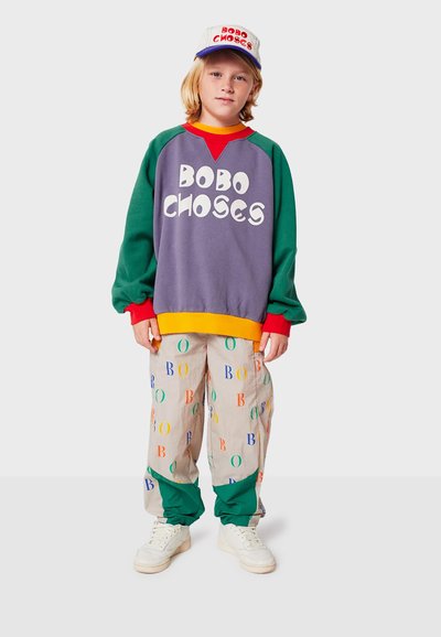 Felpa multicolore con il testo "BOBO CHOSES", maniche verdi, dettagli arancioni e rossi. Pantaloni beige chiaro con stampe colorate di lettere. Sneakers bianche.
