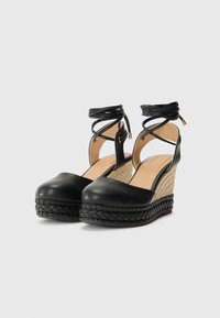 Schwarze Keilsandalen mit einem glatten Obermaterial aus Leder, einer geflochtenen Juteverbunden Plattform und überkreuzten Fesselriemen. Texturiertes schwarzes Sohle und neutrales Fußbett.