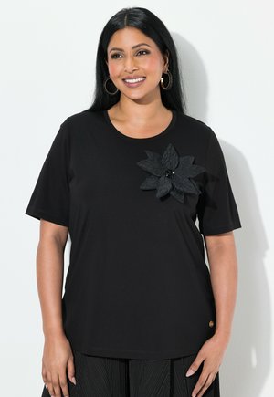 Zwart T-shirt met korte mouwen van zachte stof, voorzien van een grote zwarte bloemenversiering aan de linkerkant en een ronde halslijn.