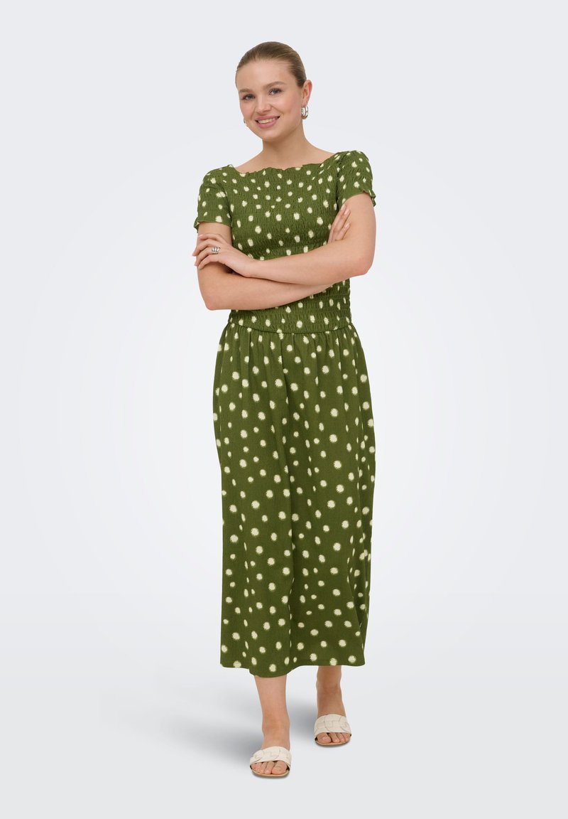 Femme souriante, les bras croisés, portant une robe midi verte à pois blancs et des sandales blanches à bout ouvert, debout sur fond blanc.