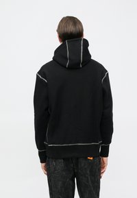 Μαύρο φούτερ με κουκούλα και ψηλό λαιμό, με έντονη ραφή, ευρύχωρη κουκούλα και τετράγωνη γραμμή. Συνδυασμένο με σκούρο παντελόνι με μοτίβο και τσέπες cargo.