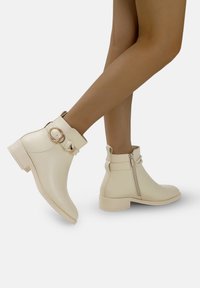 Botines en imitación cuero color crema, con un acento de hebilla redonda, cremallera lateral y tacón bajo. Textura suave con un diseño elegante.