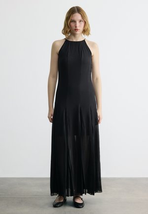 Vállig érő szőke hajú nő, ujjatlan fekete halter nyakú maxi ruhában és fekete balerina cipőben, szürke padlón állva.